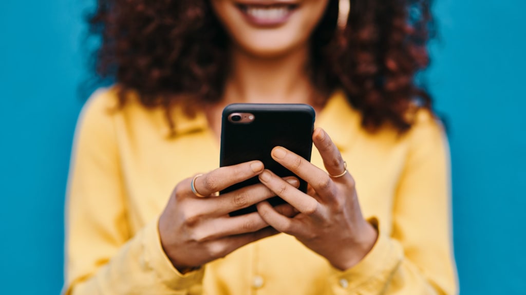 A person with curly hair wearing a yellow shirt smiles while holding and looking at a smartphone against a blue background.